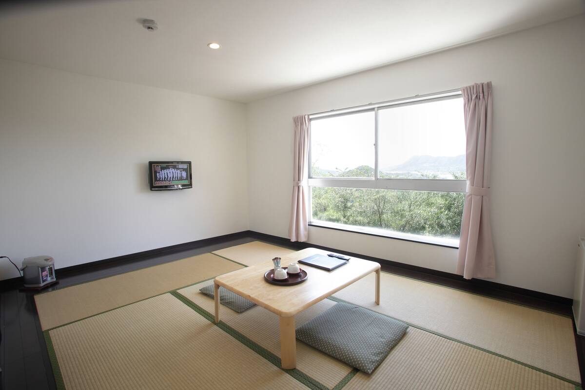 Una stanza con tatami, un tavolino in legno, un televisore a parete e una finestra con tende.