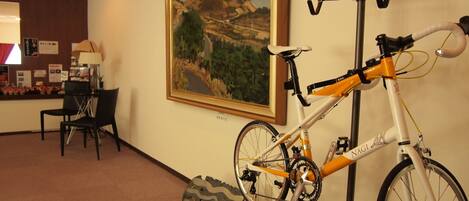 Habitación de hotel con una bicicleta colgada en la pared, un cuadro, un escritorio con una lámpara y una silla.