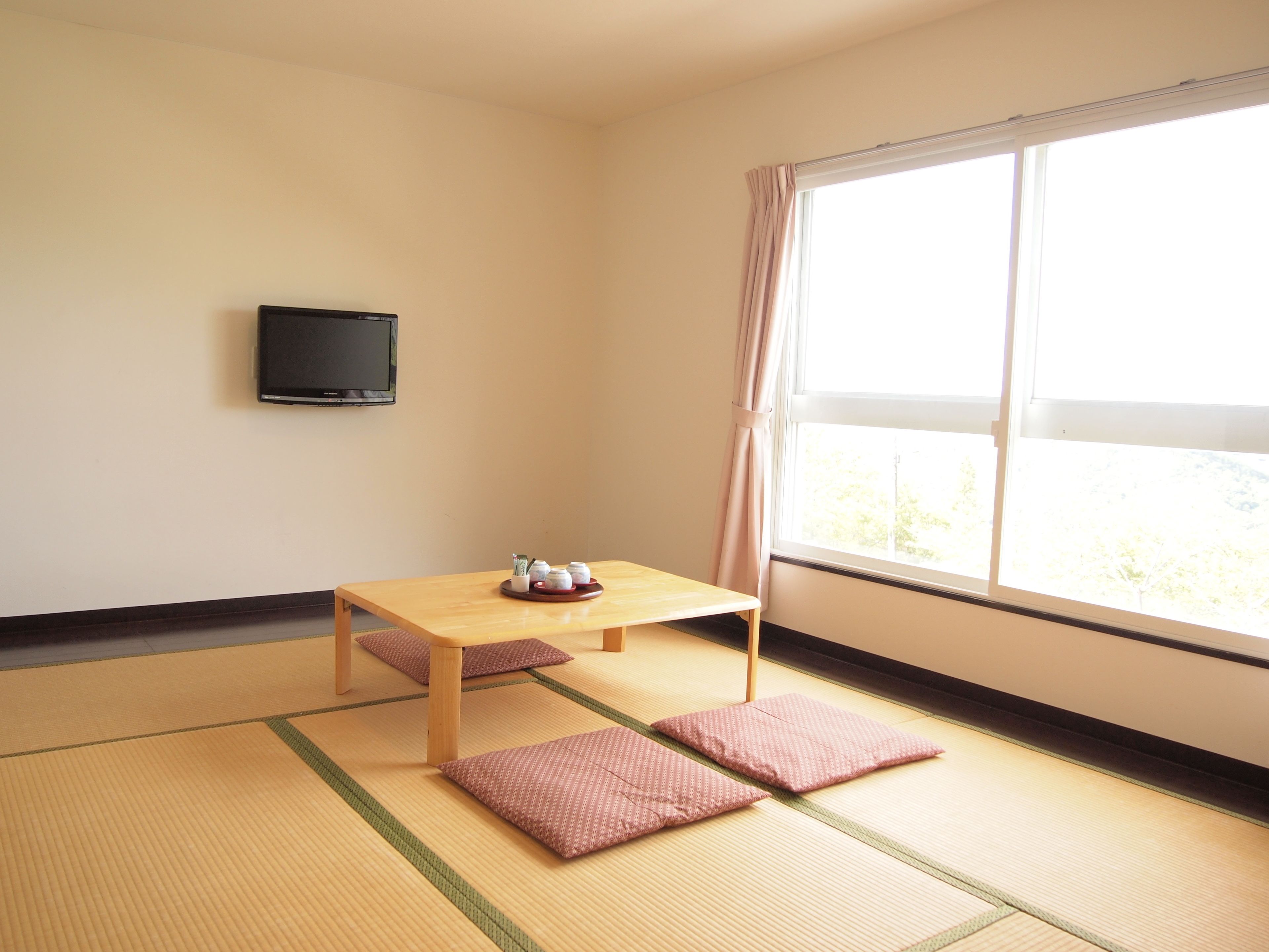 Una habitación con suelo de esteras tatami, una mesa de madera con una bandeja de objetos, un televisor en la pared y un ventanal con cortinas.