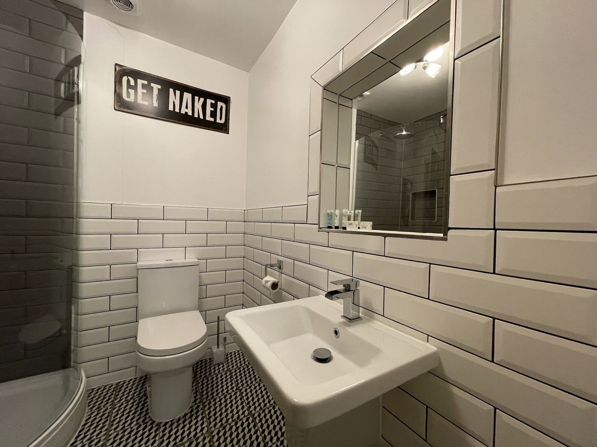 A modern bathroom with a toilet, sink, and shower. A sign on the wall reads "GET NAKED".