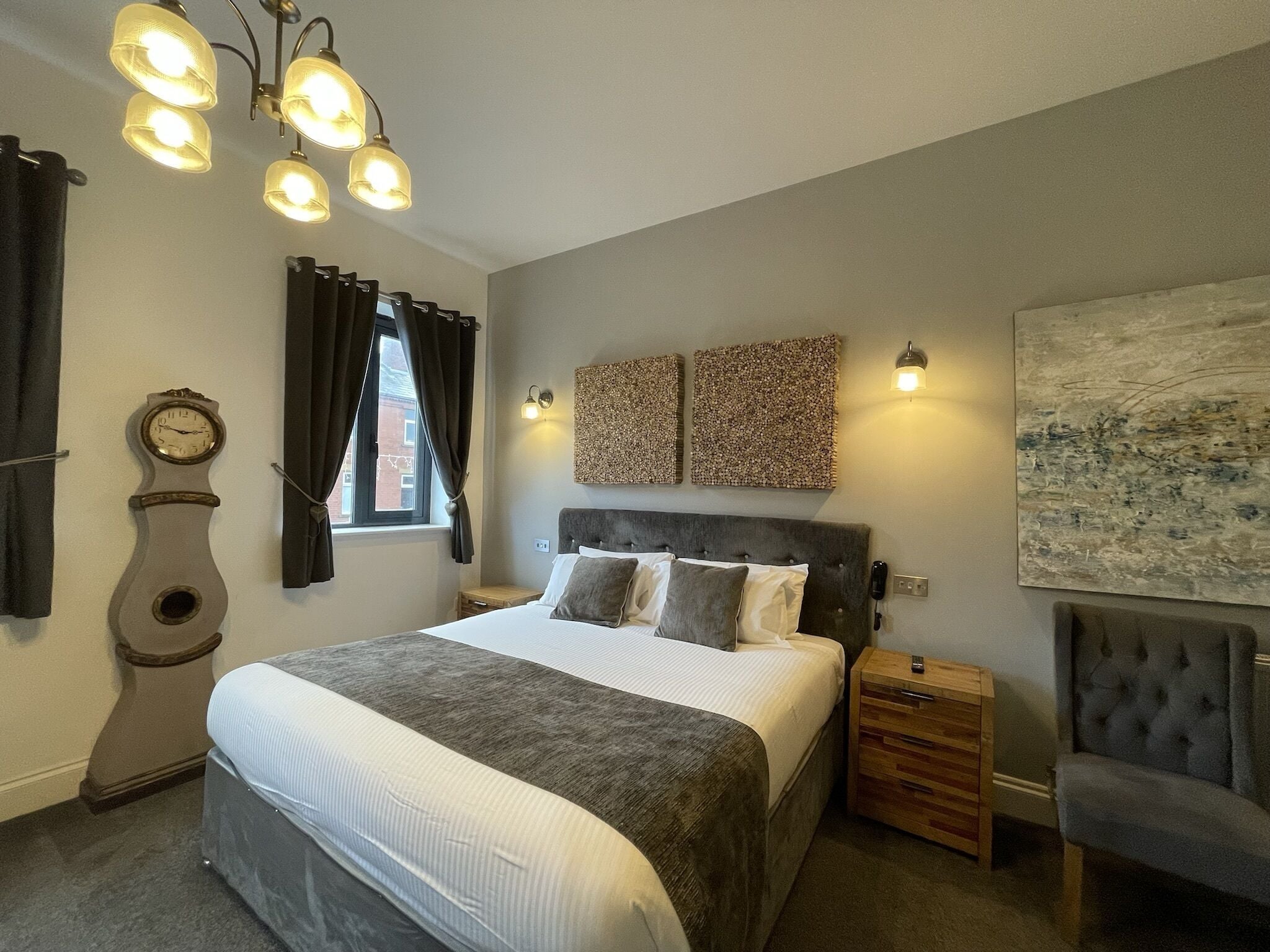 A bedroom with a large bed, a wooden bedside table, a chair, a wall clock, and a window with curtains.