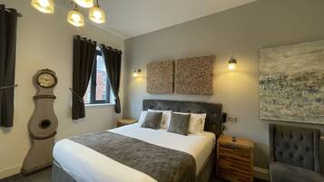 A bedroom with a large bed, a wooden bedside table, a chair, a wall clock, and a window with curtains.