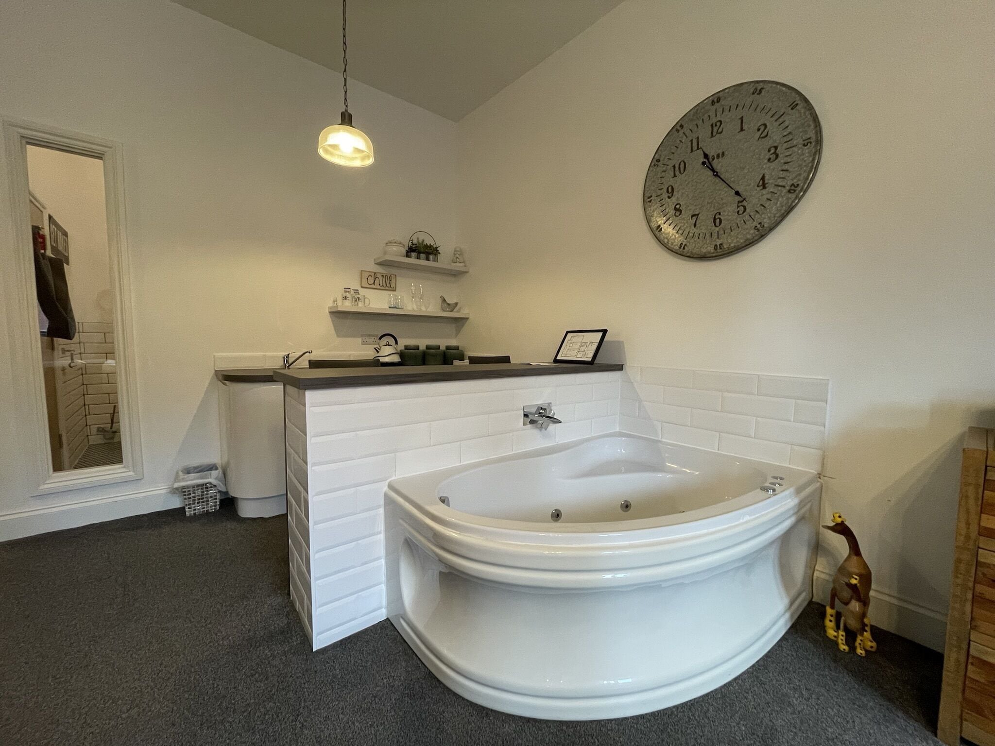 A bathroom with a white bathtub, a round wall clock, a shelf with toiletries, and a hanging light fixture.