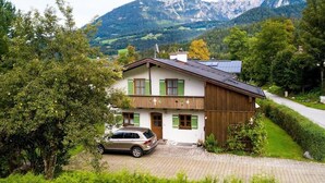 Ein zweistöckiges Haus mit einem Auto auf der Auffahrt, umgeben von viel Grün und Bergen im Hintergrund.