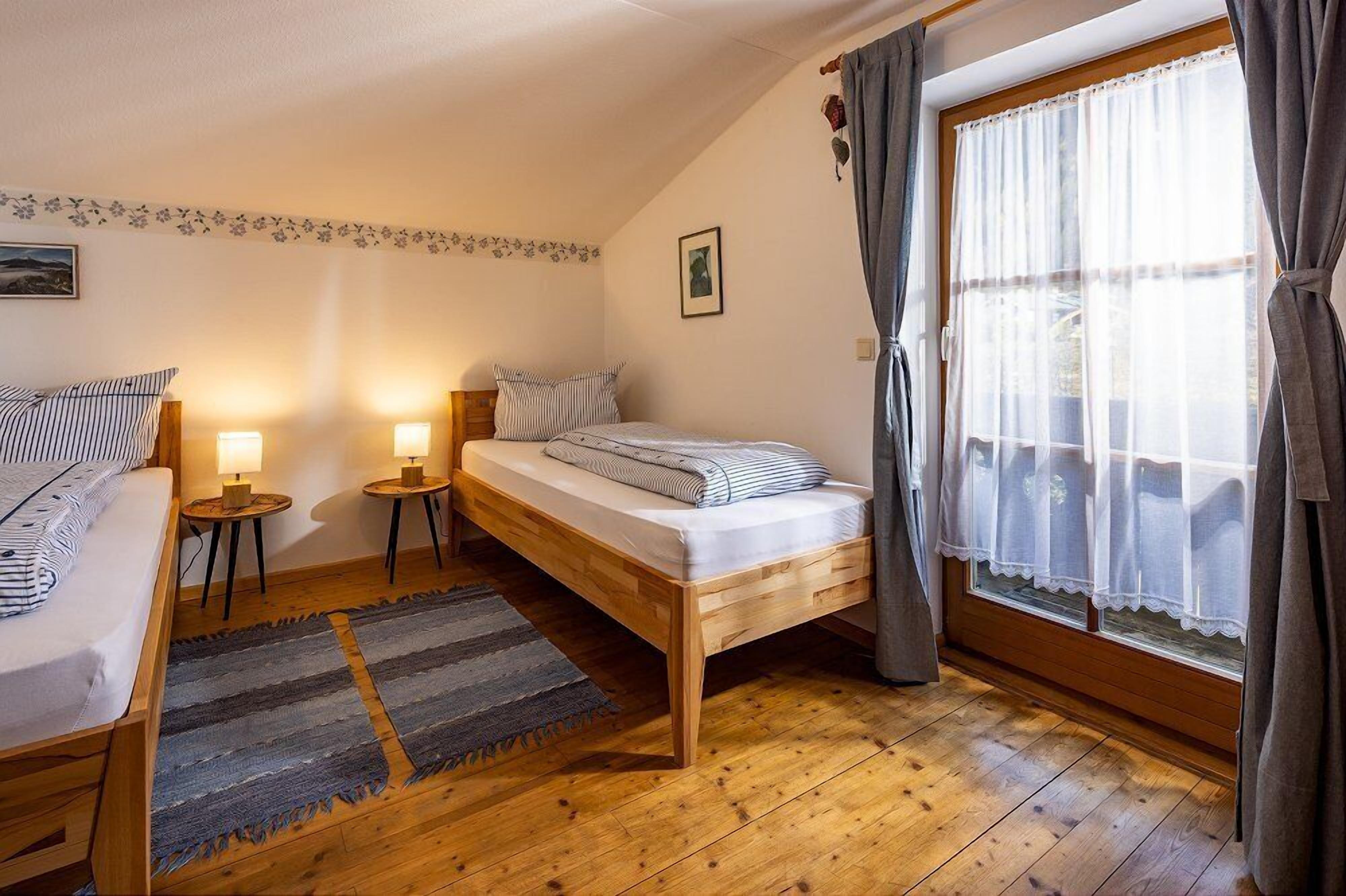 A cozy bedroom with a wooden bed, two bedside tables with lamps, a large window with curtains, and a patterned rug.