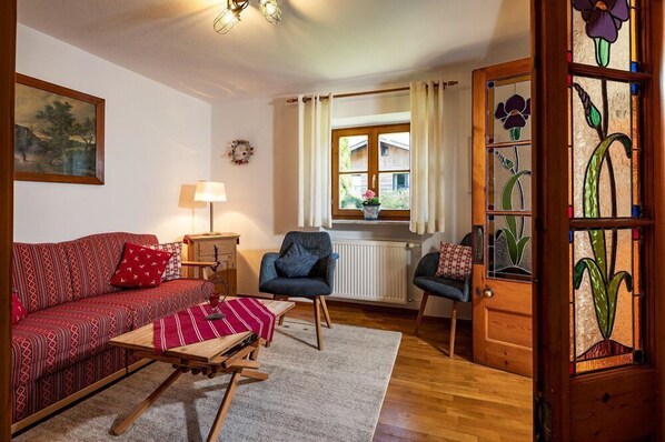 A cozy living room with a red patterned sofa, a blue armchair, a wooden coffee table, and a small table with a red and white striped tablecloth. There is a window with curtains, a radiator, and a door with stained glass.