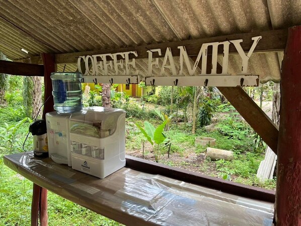 Una estación de café rústica al aire libre con un cartel que dice "COFFEE FAMILY" bajo una estructura de madera, rodeada de exuberante vegetación.