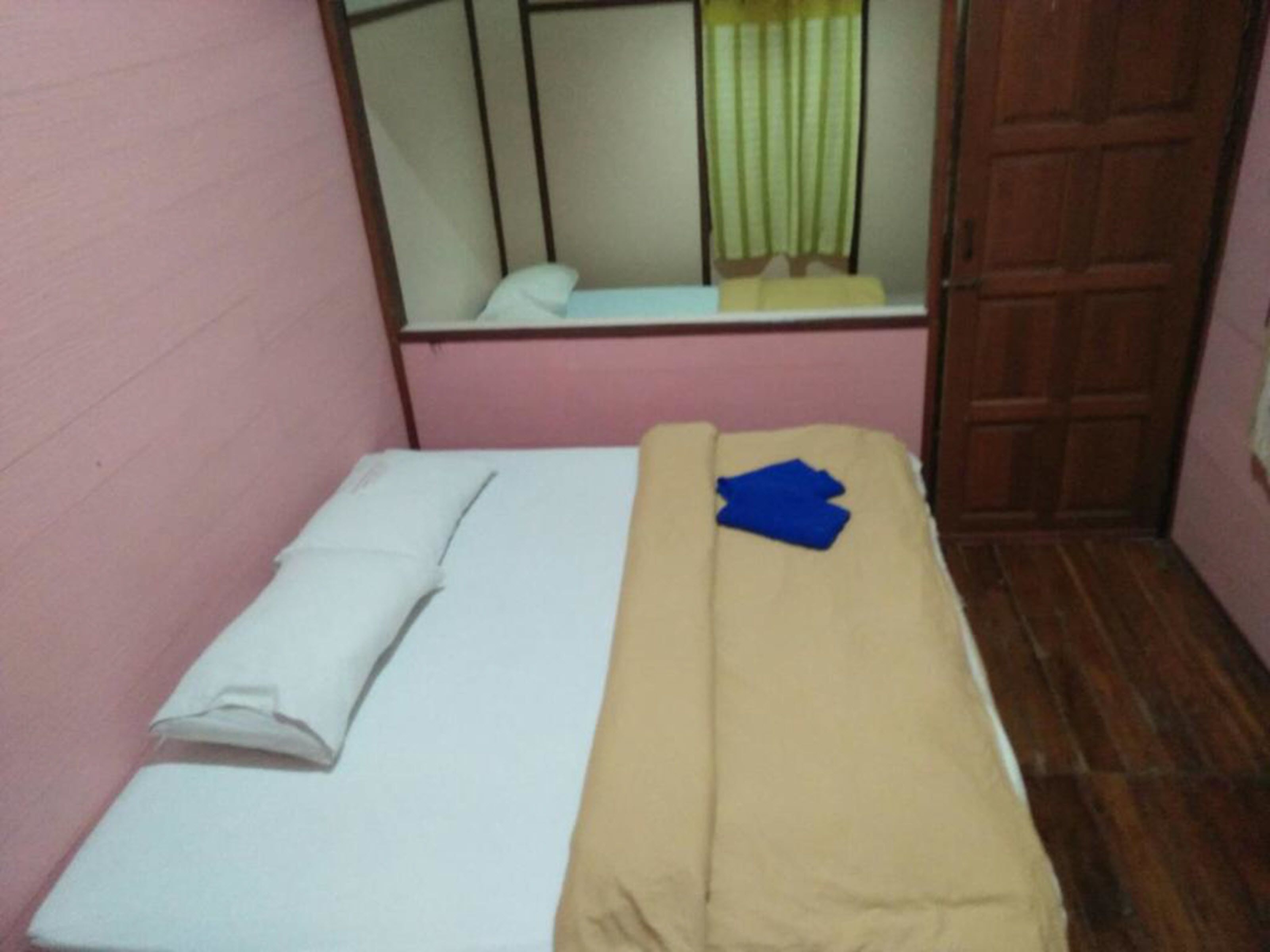 A small, single-bed room with a wooden floor, a pink wall, and a mirror reflecting a green curtain.