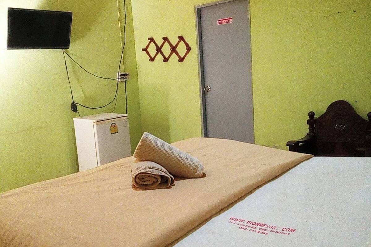 Een eenpersoonskamer met een beigekleurige matras, een opgerolde handdoek, een kleine witte verwarming en een houten hoofdbord.