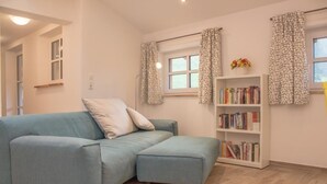 A room with a light blue sofa, a bookshelf with books, and a window with patterned curtains. - holiday flat (Siegsdorf)