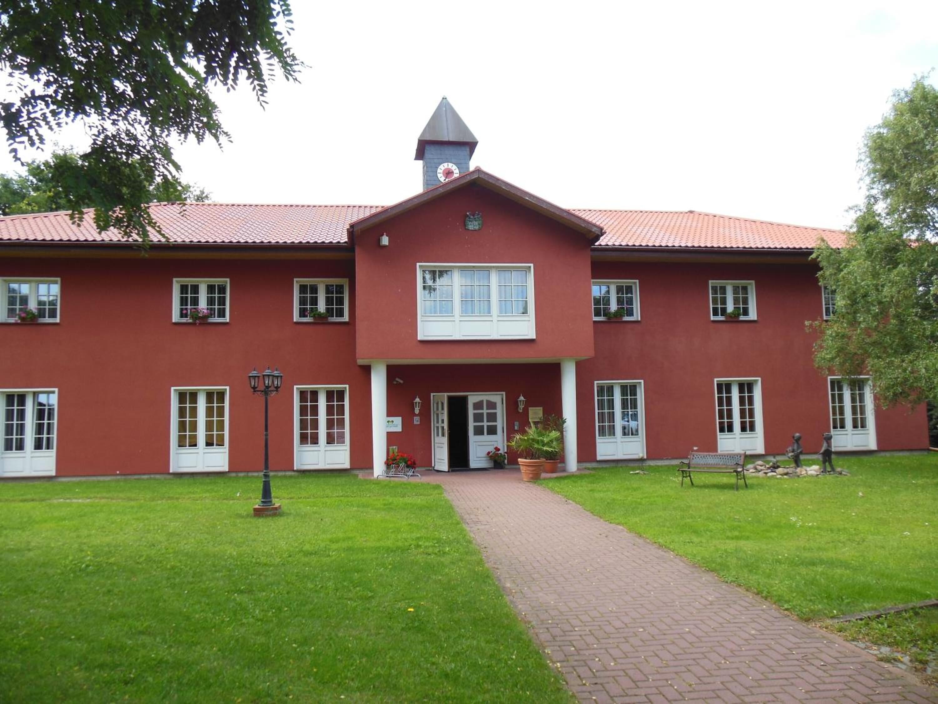 A red building with a central tower, multiple windows, and a paved pathway leading to the entrance.