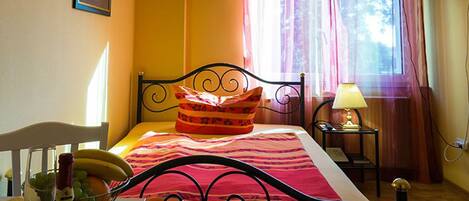 A bed with a striped bedspread, a pillow, a bedside table with a lamp, a chair, a bottle of wine, and a bowl of fruit.