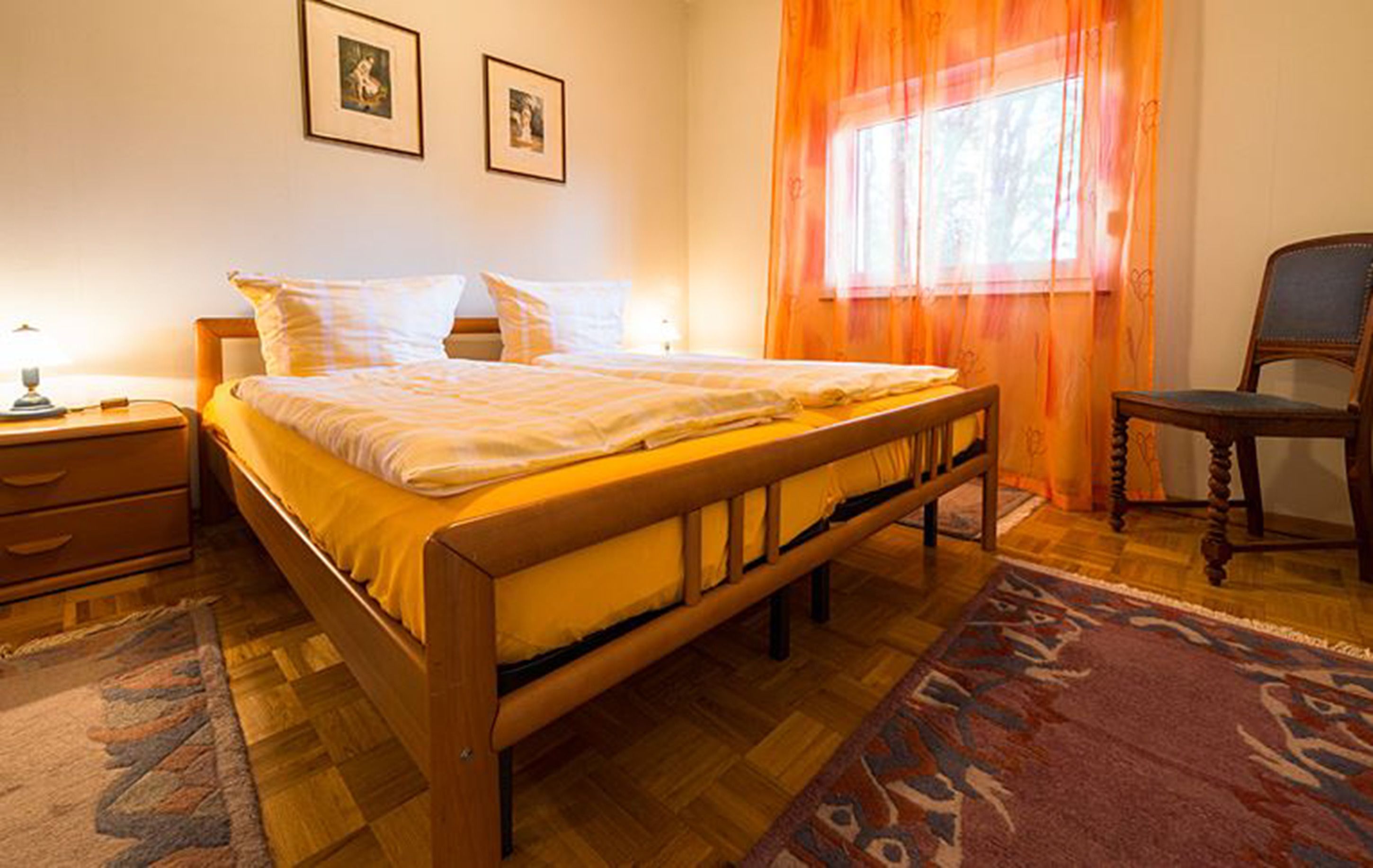 Een slaapkamer met een houten bed, twee nachtkastjes, een stoel en een raam met oranje gordijnen.