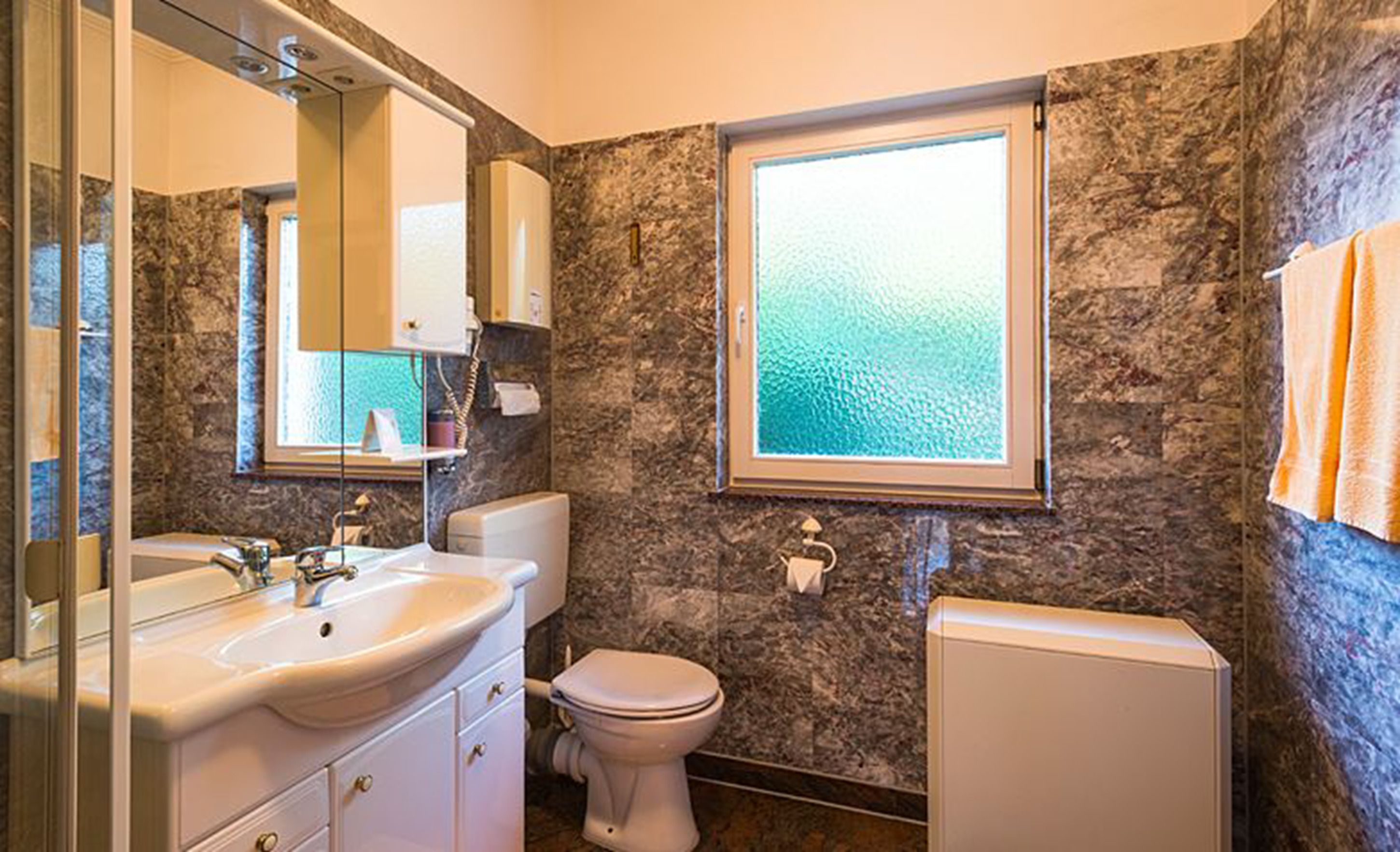A bathroom with a marble wall, a window, a sink, a toilet, and a mirror.