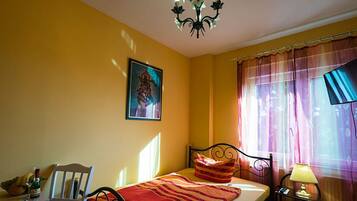 Un dormitorio con cama, una silla, una mesita de noche con lámpara, un cuadro en la pared y una ventana con cortinas.