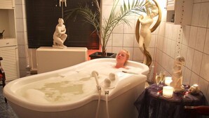 Una bañera con una persona sentada en su interior, rodeada de objetos decorativos y una pequeña mesa con velas.
