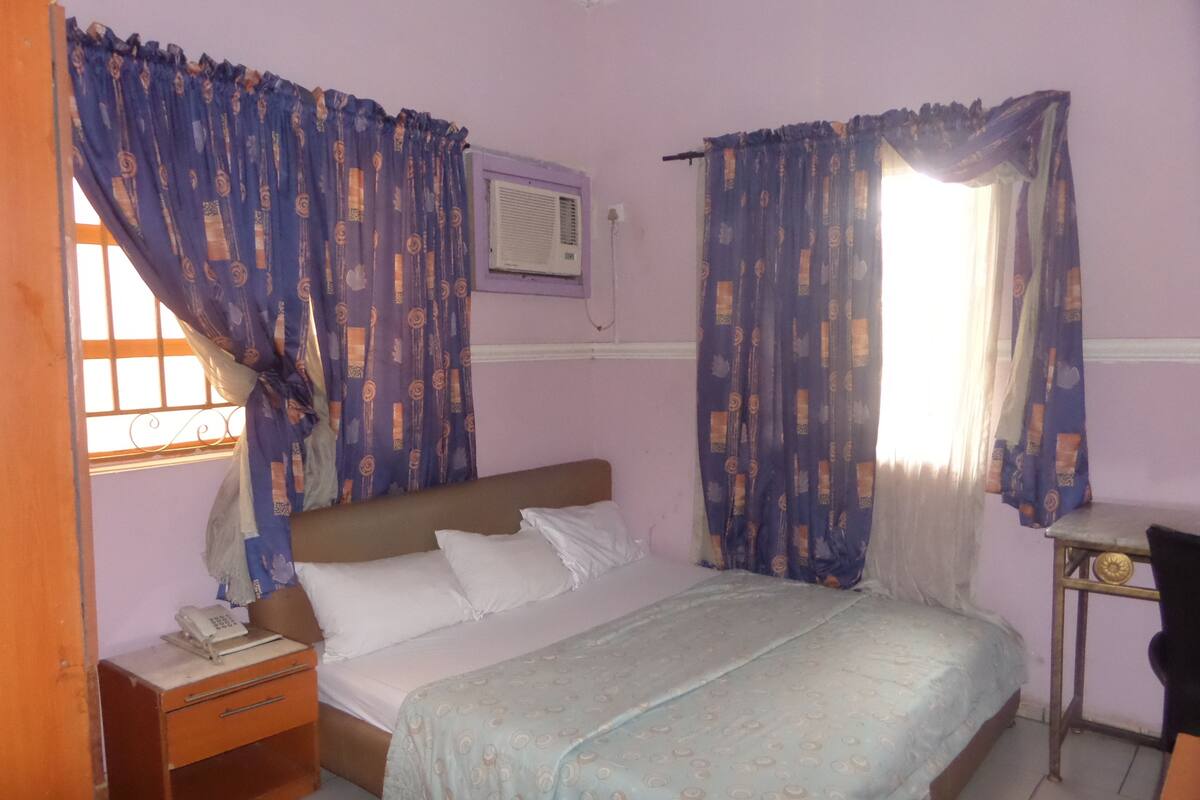 A bedroom with a bed, a nightstand, a desk, and curtains.
