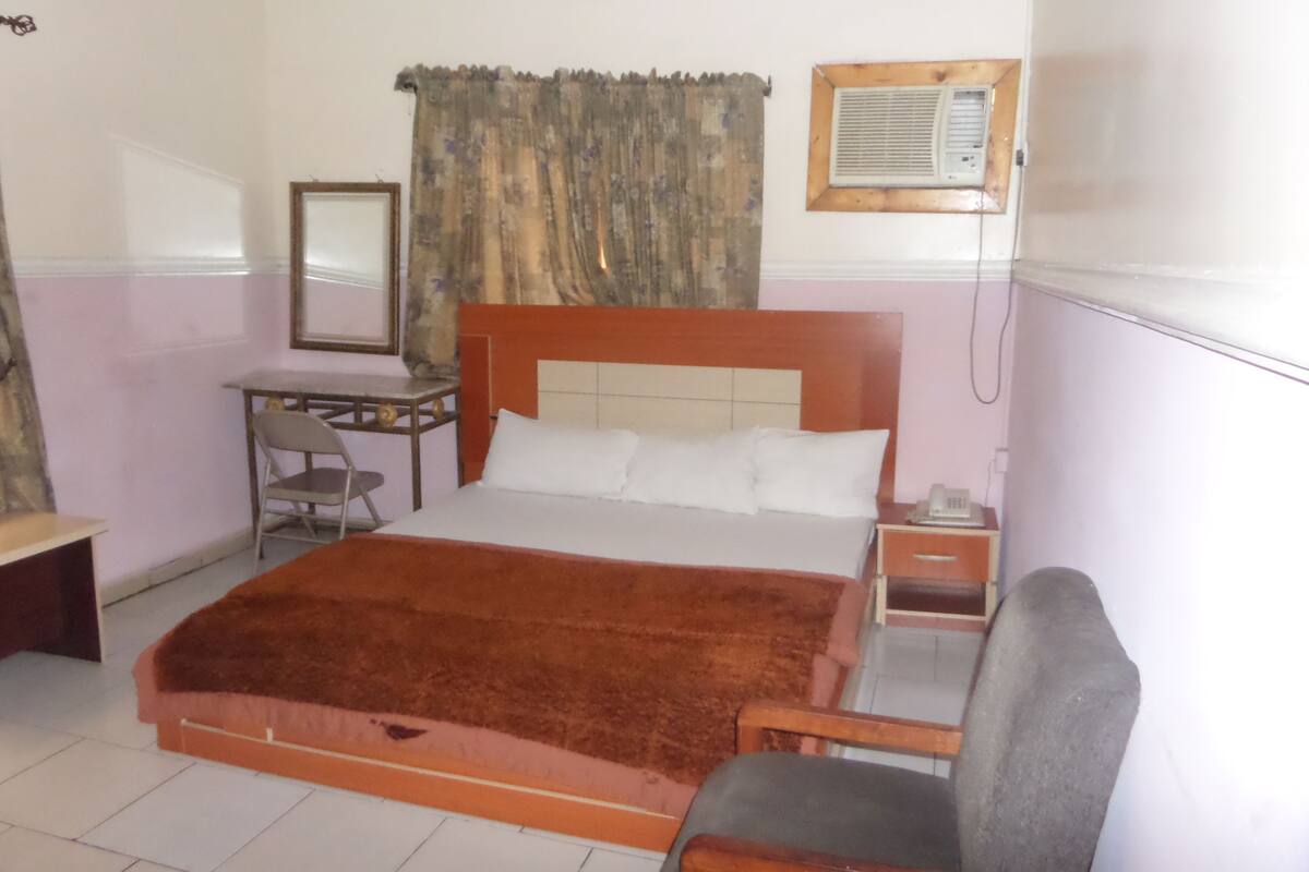 A hotel room with a bed, a desk, a chair, a nightstand, and a wall-mounted air conditioner.