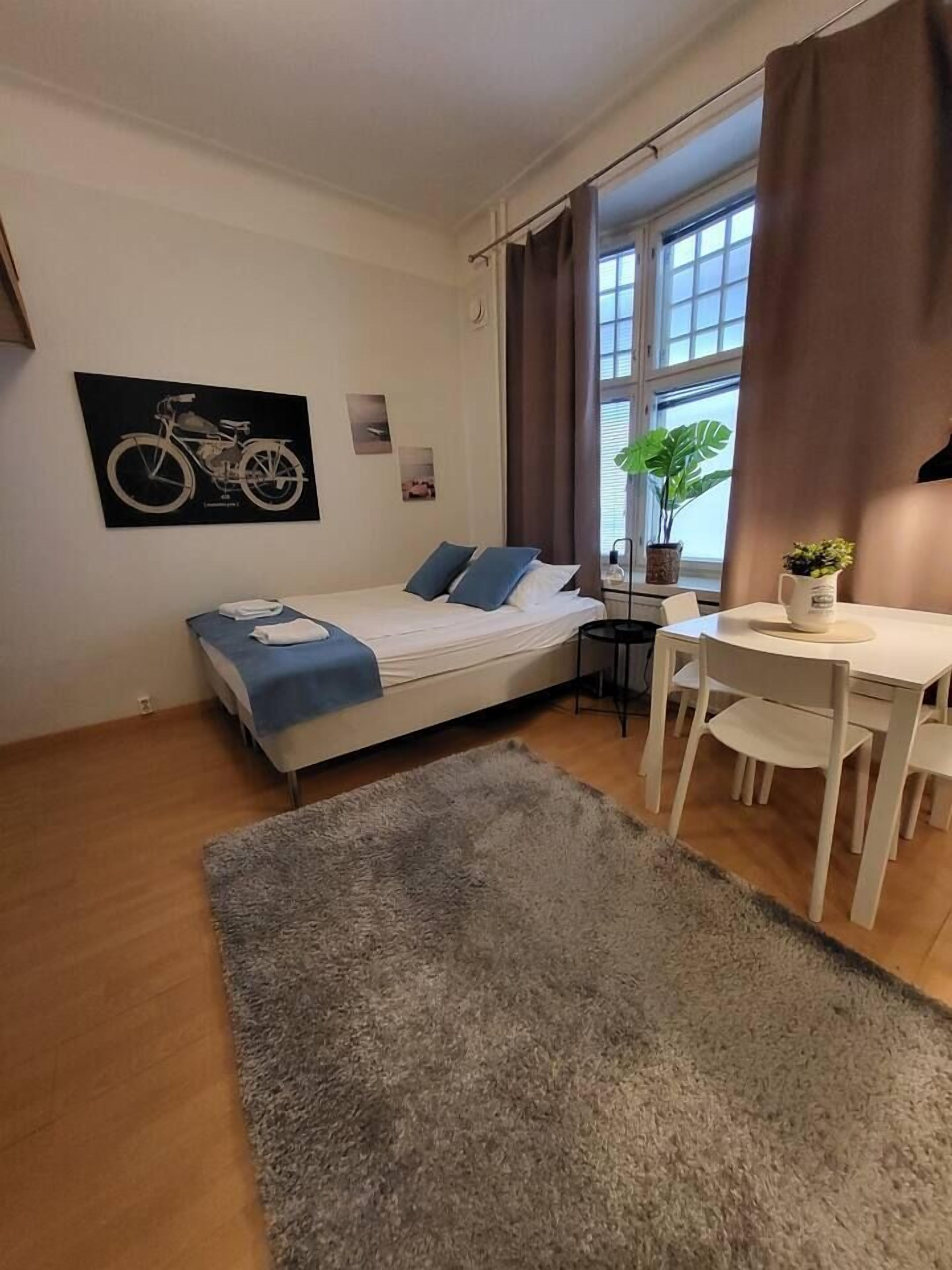 Ein Schlafzimmer mit Bett, Schreibtisch, Stuhl, einem Fenster mit Vorhängen, einer Pflanze und einem am Wand hängenden Fahrradplakat.
