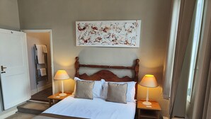 Una habitación de hotel con una cama grande, dos mesitas de noche con lámparas, un cabecero de madera, una obra de arte en la pared y una puerta a la izquierda.