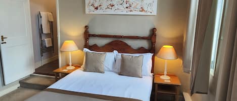 A hotel room with a large bed, two bedside tables with lamps, a wooden headboard, a wall art piece, and a door to the left.