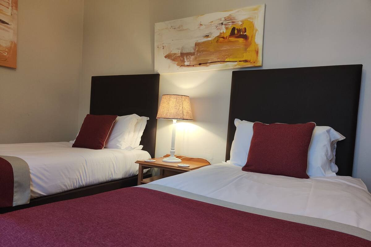 Hotellihuone, jossa on kaksi sänkyä, molemmissa valkoiset lakanat ja punaiset tyynyt, yöpöytä lampun kanssa ja taulu seinällä.