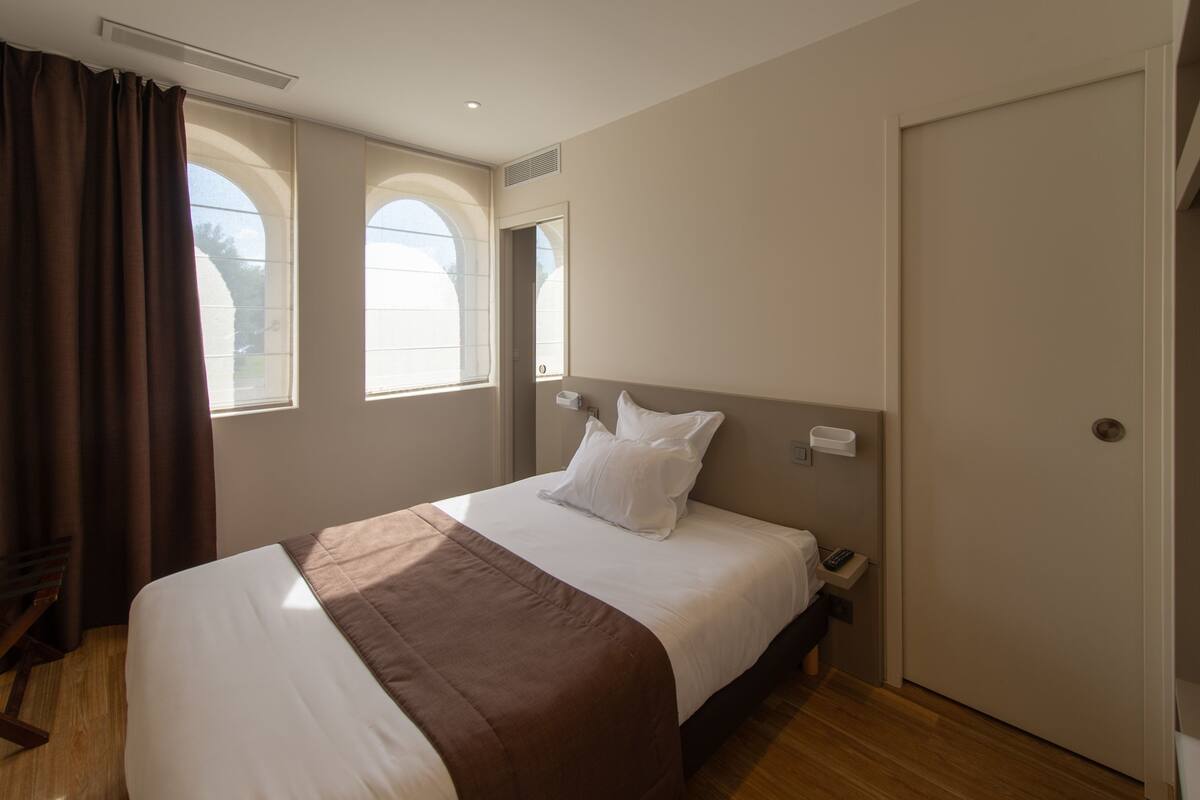 Una habitación de hotel con cama, mesita de noche, ventana con cortinas y puerta.