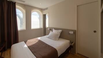 A hotel room with a bed, a bedside table, a window with curtains, and a door.