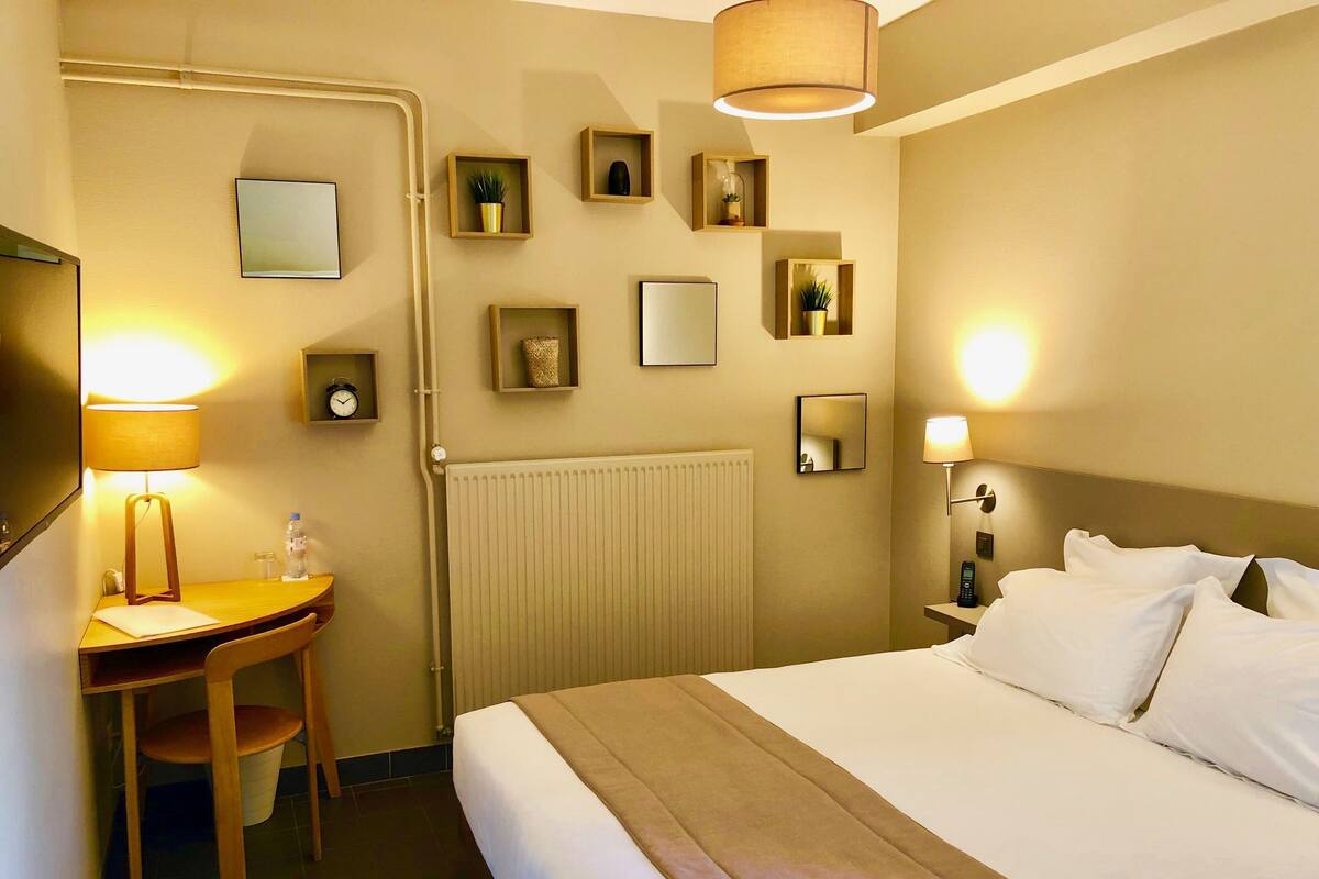 Habitación de hotel con cama, mesita de noche, lámpara, escritorio, silla y espejos en la pared.