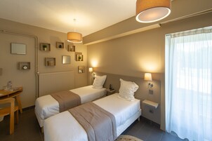 Habitación de hotel con dos camas individuales, un escritorio, una silla, una mesita pequeña, una mesita de noche, una lámpara, un espejo y una ventana con cortinas.