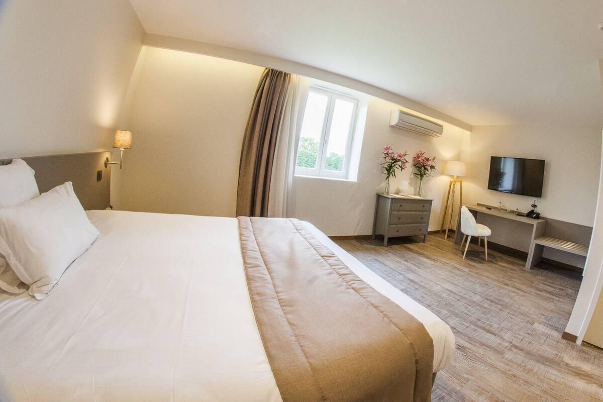 Una habitación de hotel con cama, escritorio, silla, televisión y ventana con cortinas.