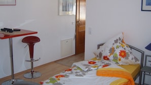 Ein kleines, ordentlich eingerichtetes Schlafzimmer mit einem Einzelbett, einem kleinen Tisch und einem Hocker.