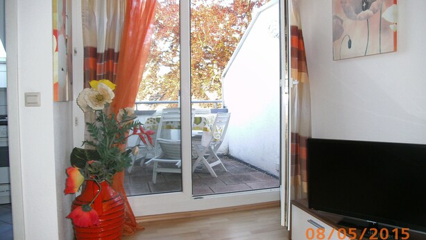 Ein Zimmer mit einer Schiebetür zu einem Außenbereich mit Tisch und Stühlen. Auf dem Boden neben der Tür steht ein roter Vasen mit Blumen.