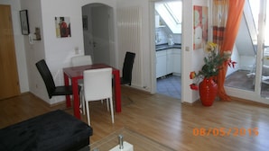 Ein modernes Wohnzimmer mit Essbereich, einer durch eine offene Tür erkennbaren Küche und einem großen Fenster mit Vorhängen.