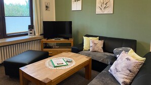 A living room with a television on a stand, a sofa, a coffee table, and a window with blinds. - Vacation apartment Schlei - Ferienhof Röhling between Schlei and Baltic Sea (Kappeln)