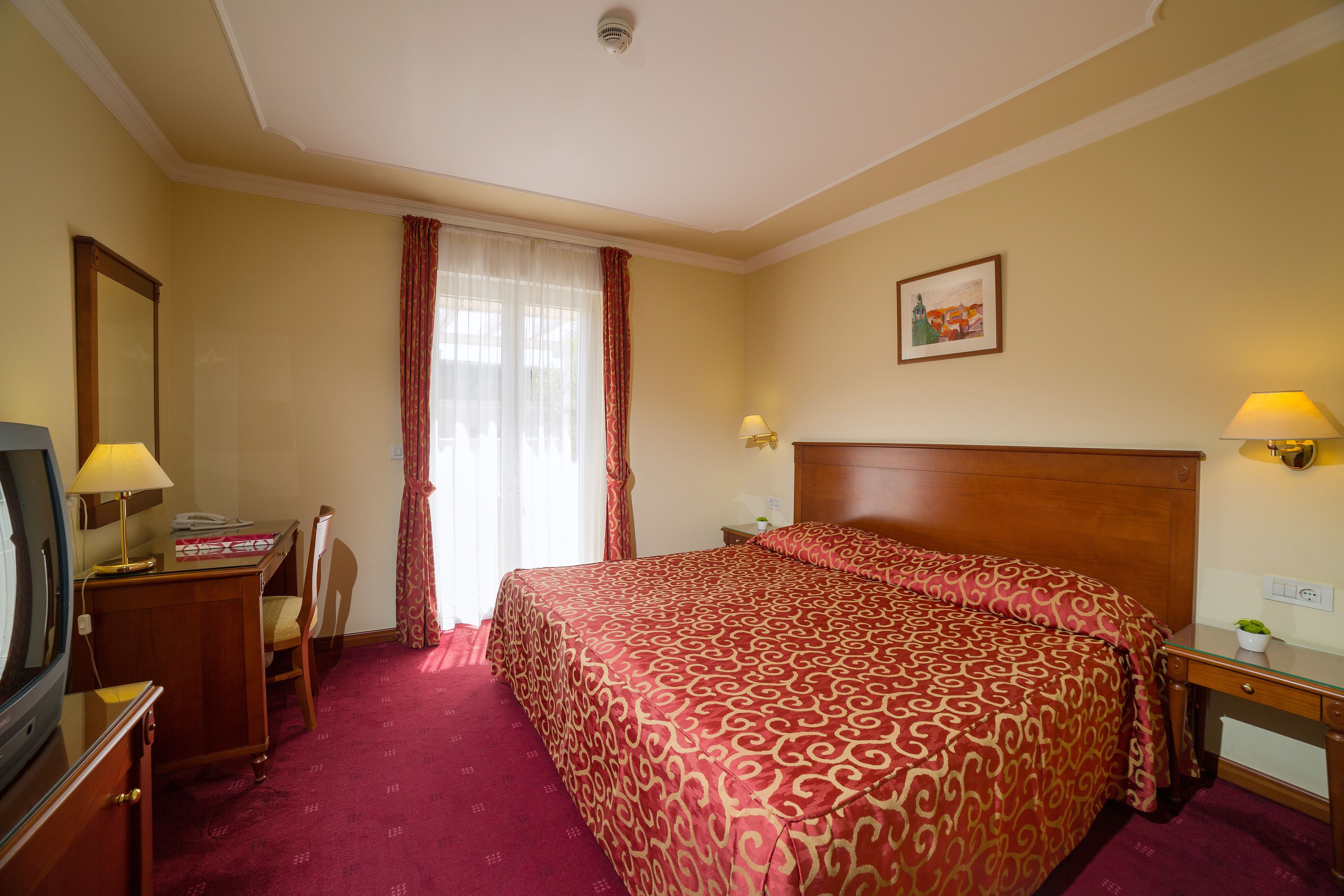 A hotel room with a large bed, a desk with a lamp, a chair, a television, and a window with curtains.