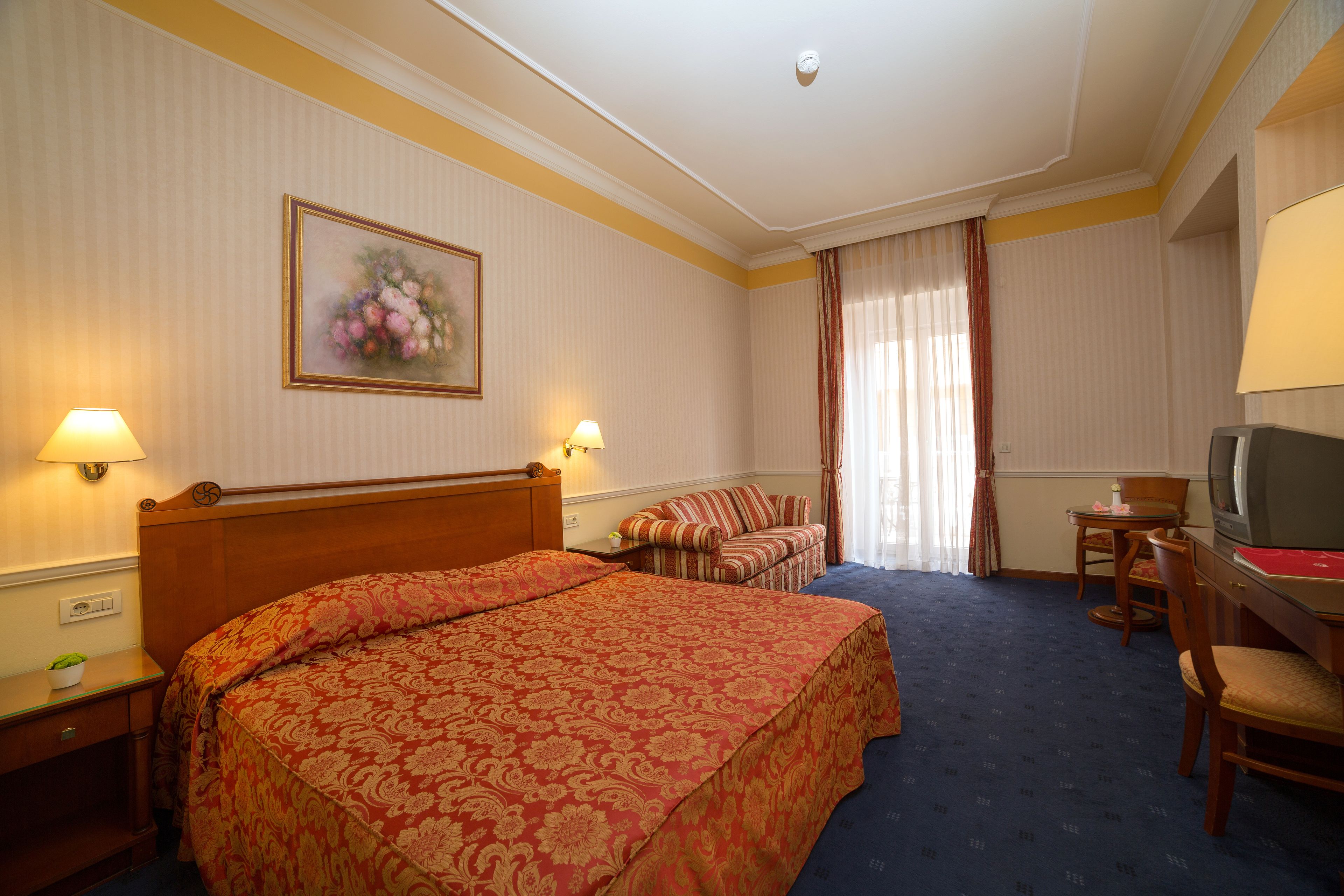 A hotel room with a large bed, a sofa, a TV, and a desk with a lamp.