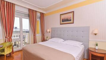 A hotel room with a large bed, a green chair, a small table, and a balcony with a view.