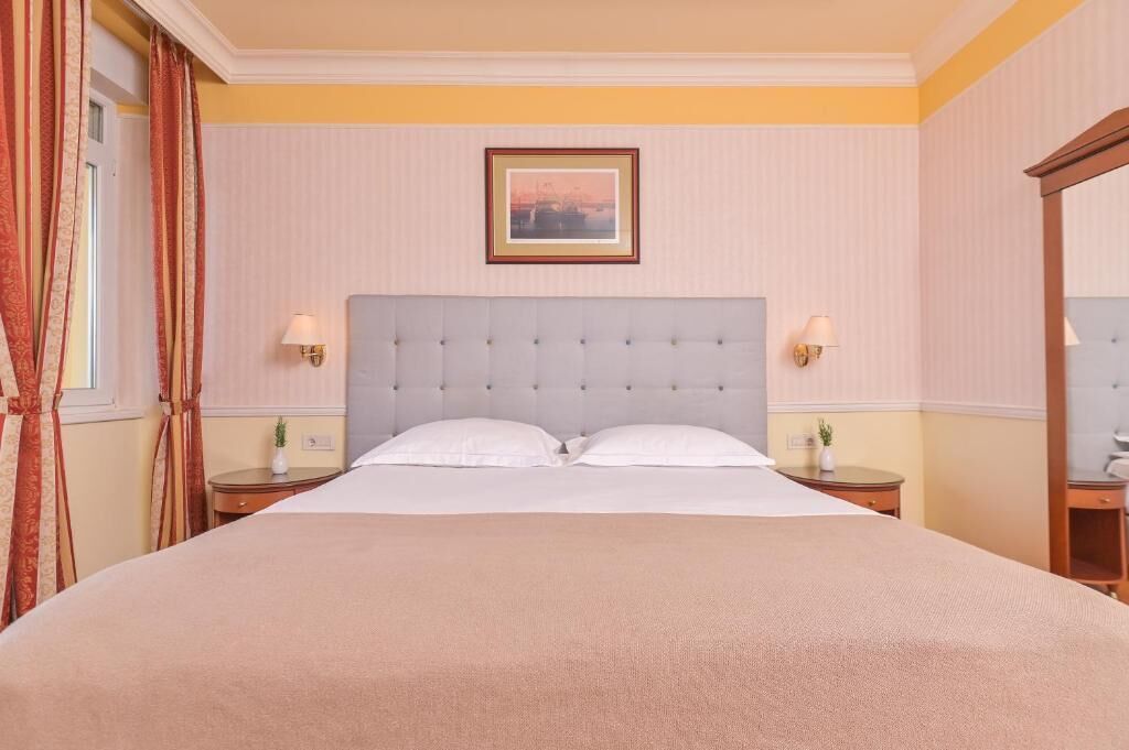 Een hotelkamer met een groot bed, twee nachtkastjes met lampen, een lijst aan de muur en een raam met gordijnen.