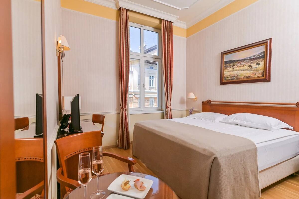 Ein Hotelzimmer mit einem Bett, einem Schreibtisch mit Computer, einem Stuhl, einem Tisch mit Speisen und zwei Weingläsern sowie einem Bild an der Wand.