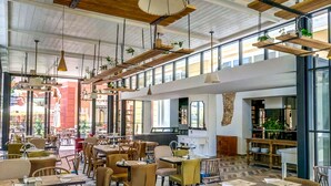 Un café spacieux avec de hauts plafonds, des poutres apparentes et des plantes suspendues. Les tables sont disposées pour accueillir les clients, et le café possède de grandes fenêtres qui offrent une vue sur la rue.