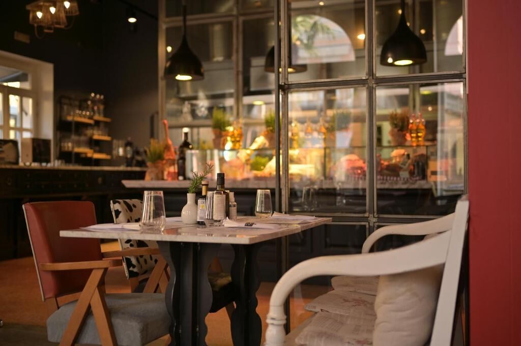 Interior de un restaurante con una mesa para dos, una zona de bar al fondo y amplias ventanas con vistas a la calle.