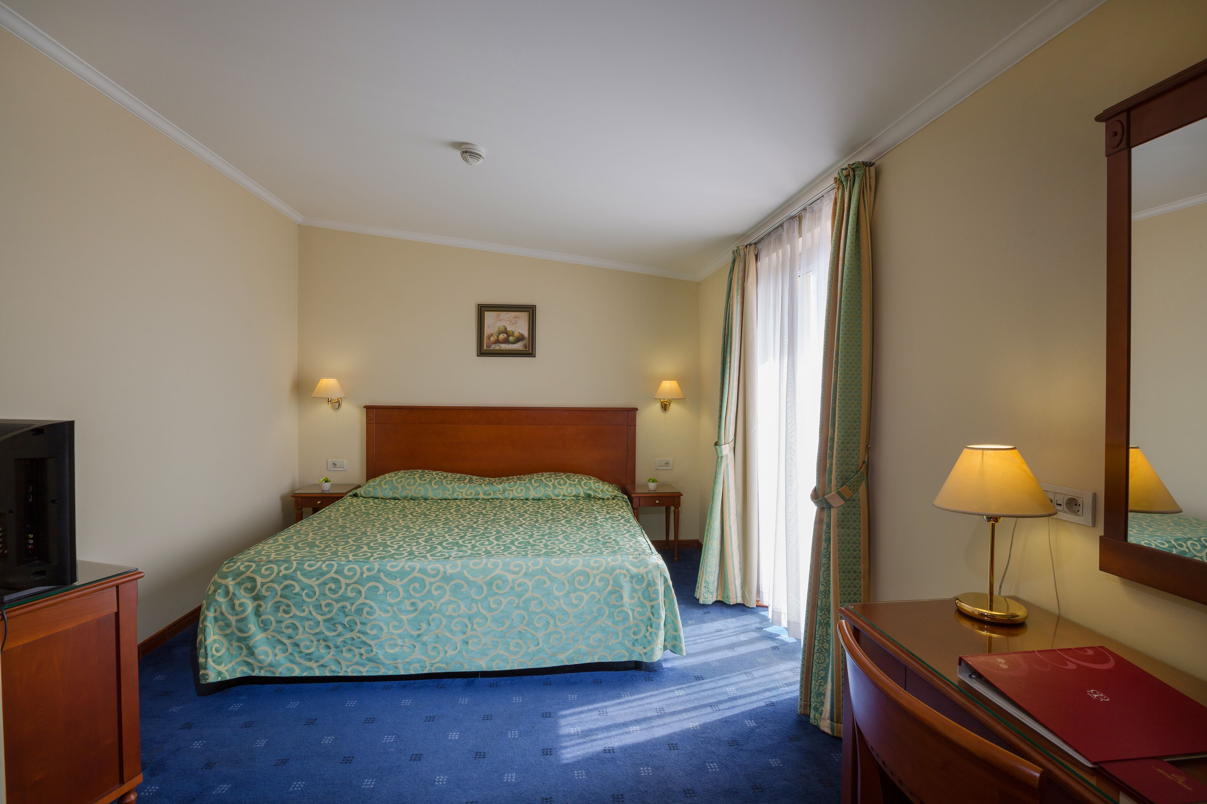 A hotel room with a bed, bedside lamps, a television, a desk with a lamp, and a mirror.