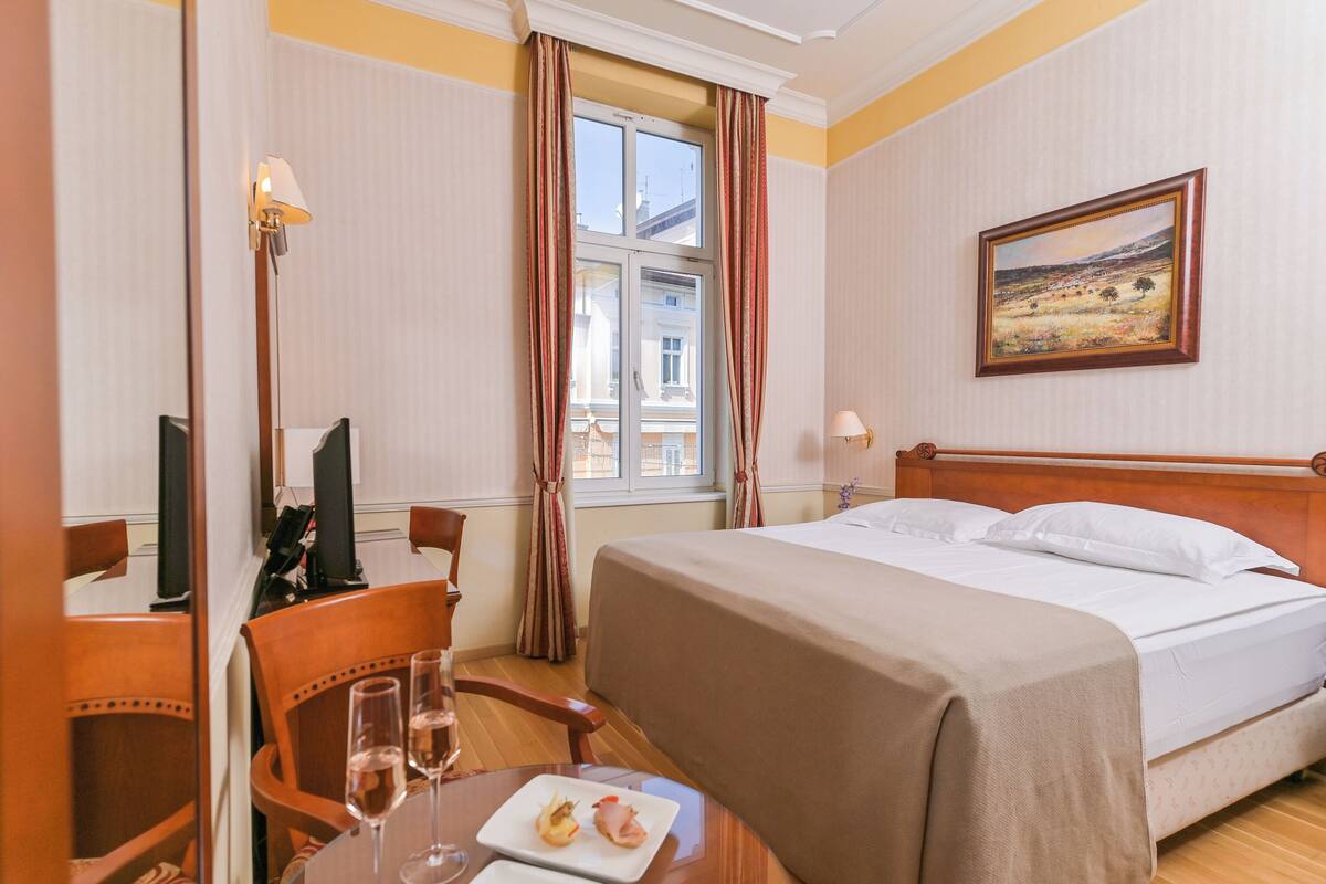 Ein Hotelzimmer mit Bett, Schreibtisch mit Stuhl, Tisch mit Teller und Gläsern sowie einem Fenster mit Vorhängen.