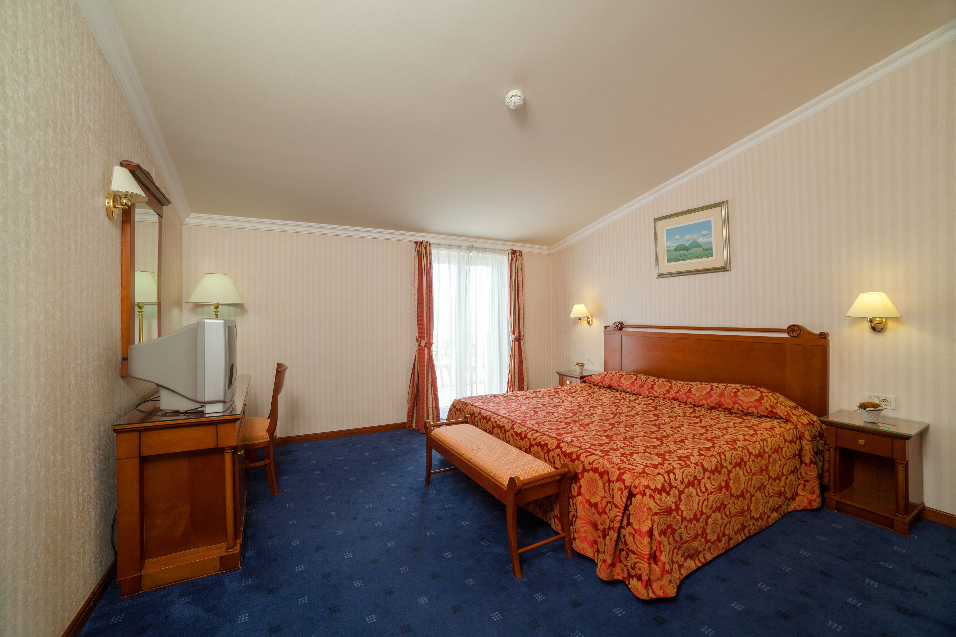 A hotel room with a bed, a desk with a computer, a chair, a bench, and two bedside lamps.