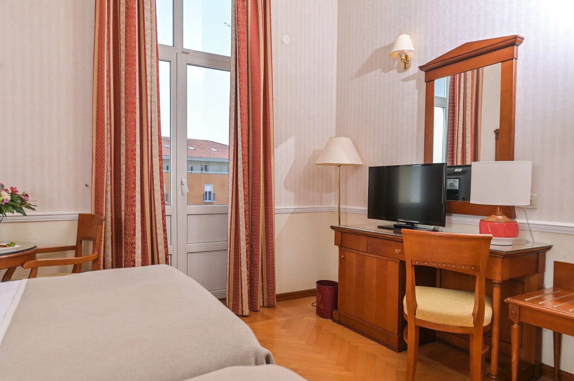 A hotel room with a bed, a desk with a television, a chair, a lamp, and a window with curtains.