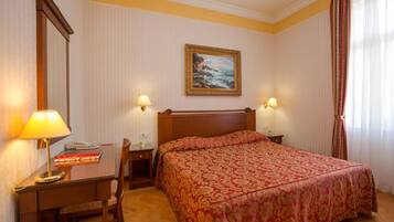 Ein Hotelzimmer mit Bett, Schreibtisch mit Lampe, Stuhl, einem Wandbild und einem Fenster mit Vorhängen.