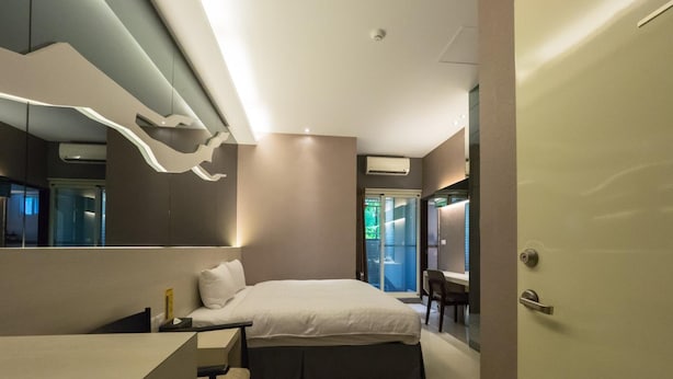 Une chambre moderne avec un lit, un bureau et une chaise. Un grand miroir est accroché au mur, et une porte se trouve sur le côté droit.