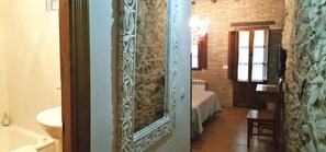 A bathroom with a white sink and toilet, a mirror with ornate frame, and a bed in the adjacent room with a brick wall and a window. - El Juacu (Llanes)