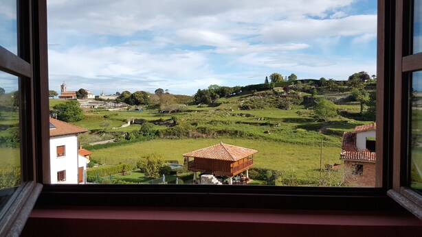 A view from a window of a rural landscape with terraced fields, a small wooden structure, and distant buildings.